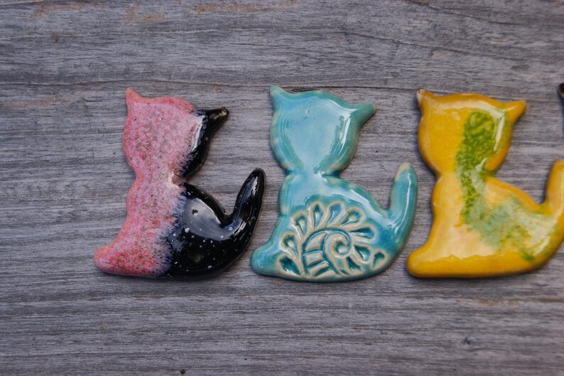 May include: Four ceramic cat figurines in different colors and designs. The cats are in a row, facing the right. The first cat is pink and black, the second is blue with a white design, the third is yellow with green accents, and the fourth is white with black accents.