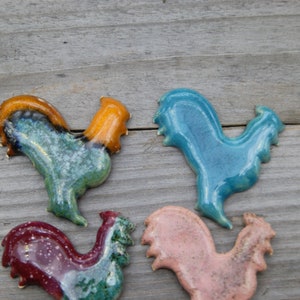 May include: Four ceramic rooster figurines in different colors. The roosters are blue, green, pink, and red with brown accents.