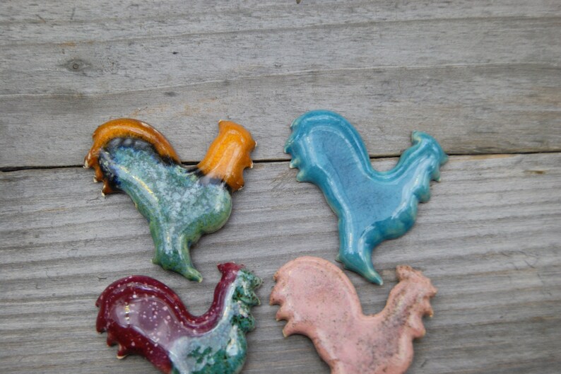 May include: Four ceramic rooster figurines in different colors. The roosters are blue, green, pink, and red with brown accents.