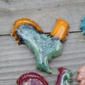 May include: A collection of ceramic rooster figurines in various colors, including blue, green, orange, and pink. The roosters are hand-painted and have a rustic, vintage look.