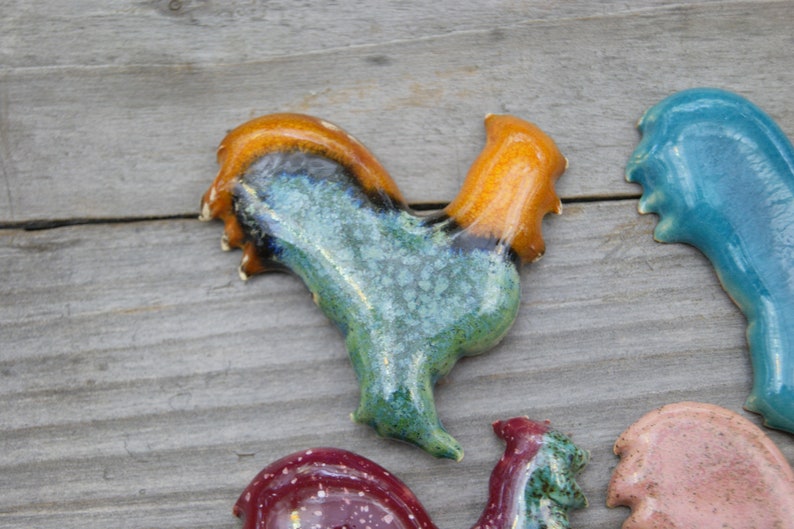 May include: A collection of ceramic rooster figurines in various colors, including blue, green, orange, and pink. The roosters are hand-painted and have a rustic, vintage look.