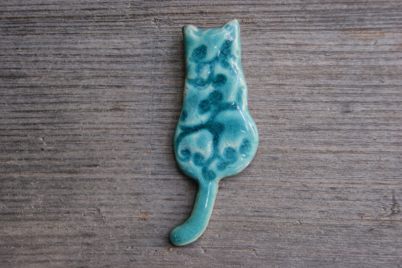 May include: A teal blue ceramic cat figurine with a white floral design. The cat is sitting with its tail curled around its body.