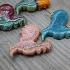 May include: Four ceramic figurines in the shape of a bird, each painted in a different color: green, blue, pink, and orange. The figurines are arranged on a wooden surface.
