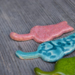May include: Three ceramic cat figurines in different colors and patterns. The first cat is pink with a speckled finish, the second is blue with a swirling pattern, and the third is green with a yellow stripe.