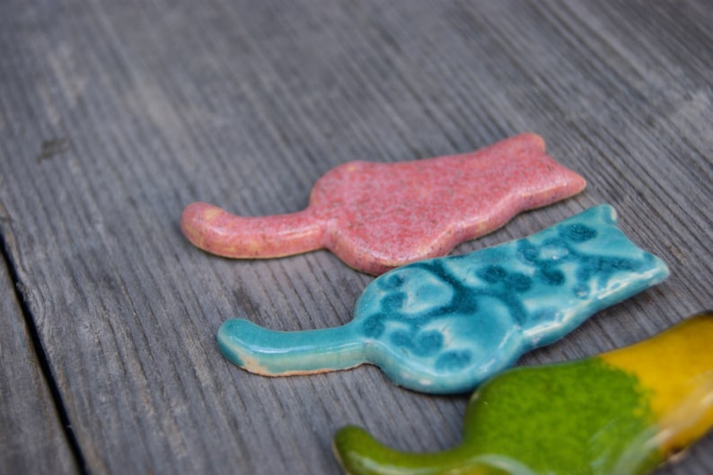 May include: Three ceramic cat figurines in different colors and patterns. The first cat is pink with a speckled finish, the second is blue with a swirling pattern, and the third is green with a yellow stripe.