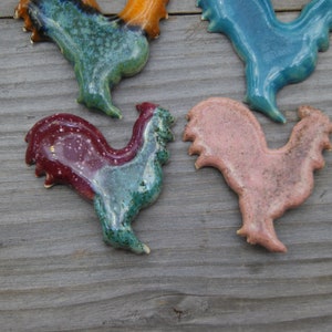 May include: Four ceramic rooster figurines in different colors. The roosters are in various poses, with one standing upright, one with its head tilted, and two with their wings spread. The roosters are painted in shades of blue, green, pink, and red.
