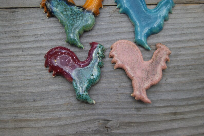 May include: Four ceramic rooster figurines in different colors. The roosters are in various poses, with one standing upright, one with its head tilted, and two with their wings spread. The roosters are painted in shades of blue, green, pink, and red.