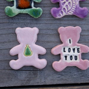 May include: Two pink ceramic bear figurines. One bear has a green pear design on its chest. The other bear has the words "I LOVE YOU" spelled out on its chest.