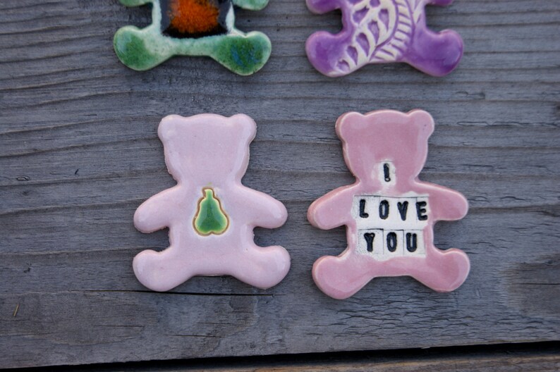 May include: Two pink ceramic bear figurines. One bear has a green pear design on its chest. The other bear has the words "I LOVE YOU" spelled out on its chest.