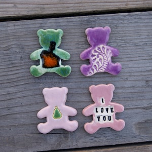 May include: Four ceramic bear figurines, two pink, one green, and one purple. The pink bears have different designs, one with a pear and the other with the words "I Love You". The green bear has a brown and orange design, and the purple bear has a white floral design.
