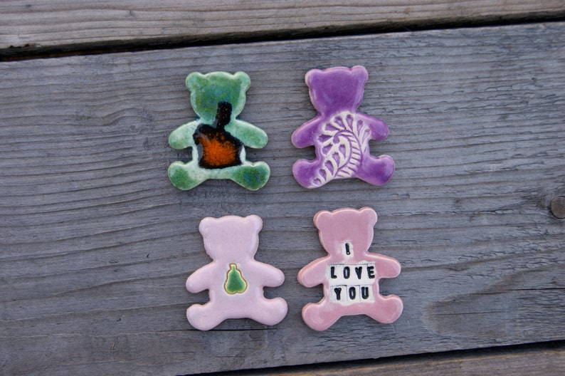 May include: Four ceramic bear figurines, two pink, one green, and one purple. The pink bears have different designs, one with a pear and the other with the words "I Love You". The green bear has a brown and orange design, and the purple bear has a white floral design.