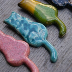 May include: Four ceramic cat figurines in different colors: pink, blue, green, and black. The cats are all in a similar pose, with their tails curled around their bodies.