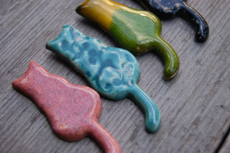 May include: Four ceramic cat figurines in different colors: pink, blue, green, and black. The cats are all in a similar pose, with their tails curled around their bodies.
