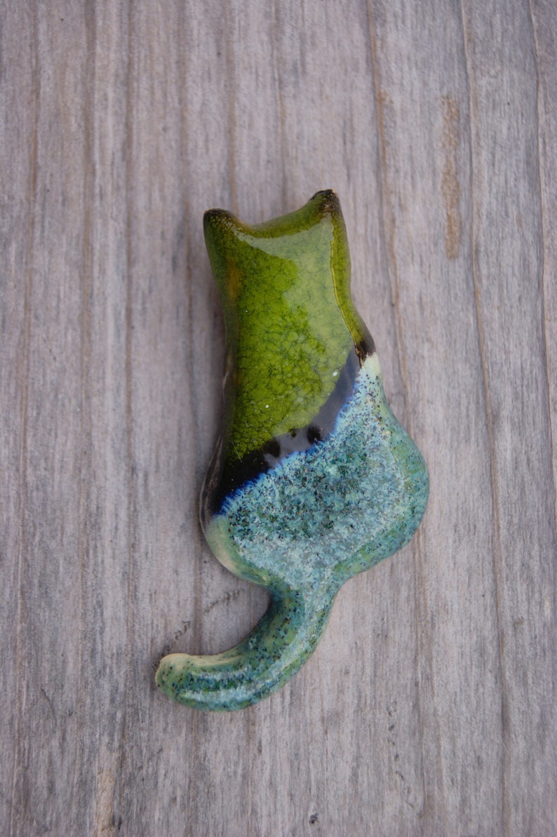 May include: A ceramic cat figurine with a green, blue, and black glaze. The cat is sitting with its tail curled around its body.