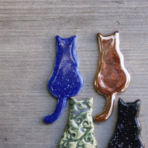 May include: Four ceramic cat figurines in different colors and patterns. The cats are facing away from the viewer and have a simple, minimalist design.