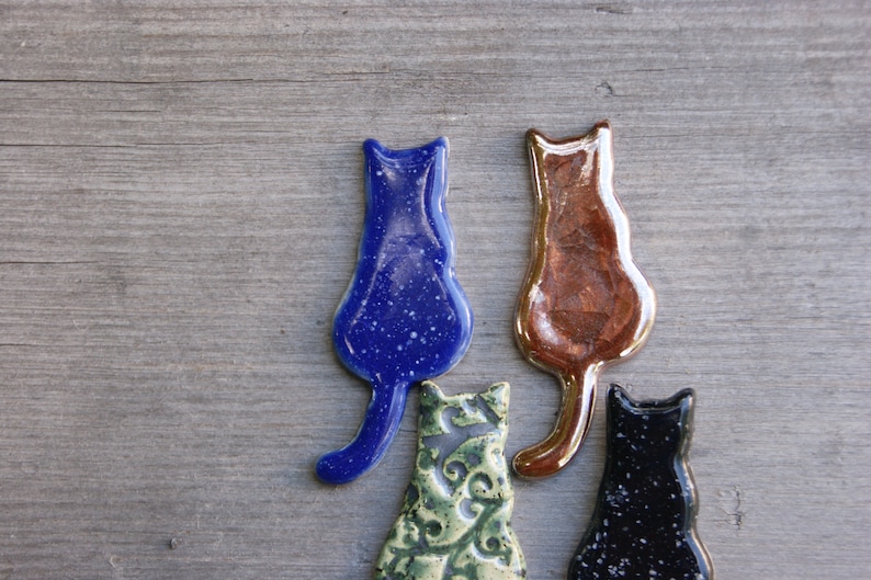 May include: Four ceramic cat figurines in different colors and patterns. The cats are facing away from the viewer and have a simple, minimalist design.