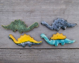 Może przedstawiać: Cztery ceramiczne figurki dinozaurów w odcieniach zieleni, niebieskiego, żółtego i szarego. Każda figurka ma teksturowane, szkliwione wykończenie i jest umieszczona na tle zwietrzałego drewna. Dinozaury są w różnych pozach.