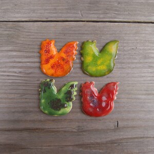 Op de afbeelding: Vier keramische haanfiguren in verschillende kleuren: oranje met rode stippen, groen met bruine stippen, groen met gele stippen en rood met zilveren stippen.