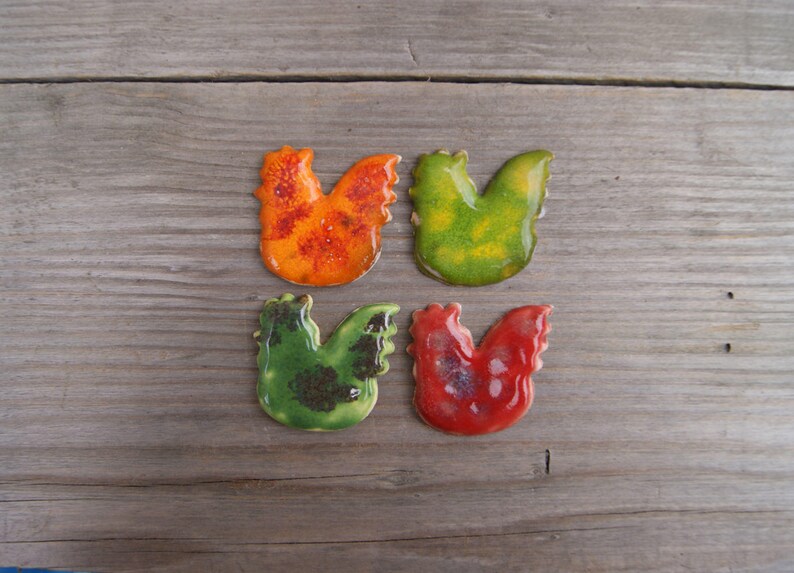 Op de afbeelding: Vier keramische haanfiguren in verschillende kleuren: oranje met rode stippen, groen met bruine stippen, groen met gele stippen en rood met zilveren stippen.
