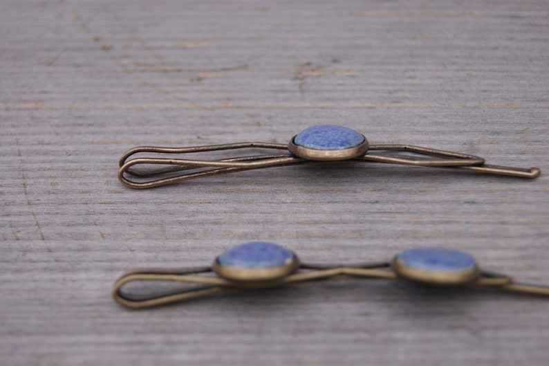 Puede incluir: Dos pinzas para el pelo doradas con detalles de piedras preciosas azules. Las pinzas son sencillas y elegantes, perfectas para a&ntilde;adir un toque de brillo a cualquier peinado.