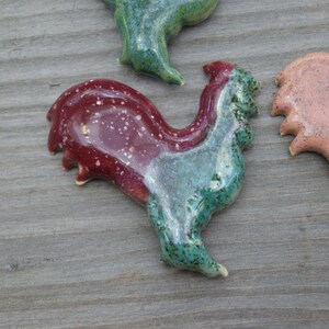May include: A ceramic rooster figurine with a red and green glaze. The rooster is standing on a wooden surface.