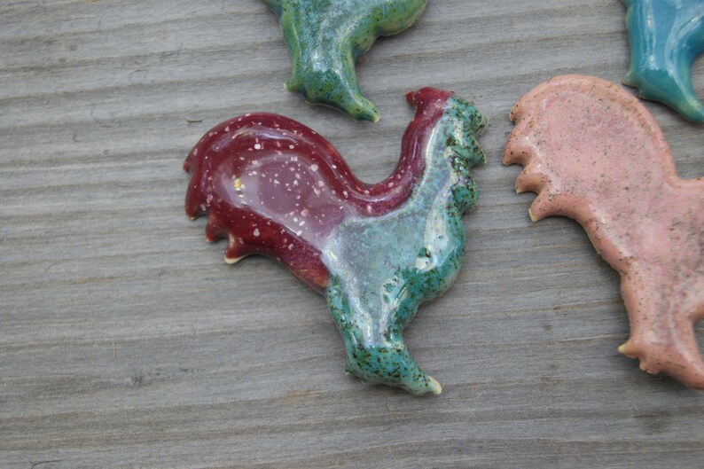 May include: A ceramic rooster figurine with a red and green glaze. The rooster is standing on a wooden surface.
