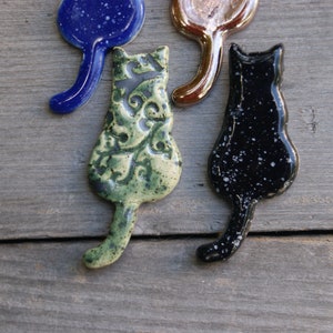 May include: Four ceramic cat figurines in different colors and patterns. The cats are all facing the same direction with their tails curled up behind them. The cats are green with a floral pattern, black with white speckles, blue with white speckles, and brown with a metallic sheen.