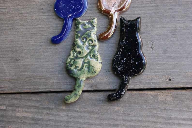 May include: Four ceramic cat figurines in different colors and patterns. The cats are all facing the same direction with their tails curled up behind them. The cats are green with a floral pattern, black with white speckles, blue with white speckles, and brown with a metallic sheen.