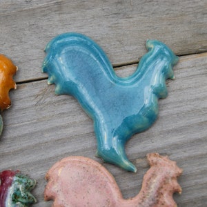 May include: A collection of ceramic rooster figurines in various colors, including teal, orange, pink, and green. The roosters are all in a standing position with their heads tilted to the side.