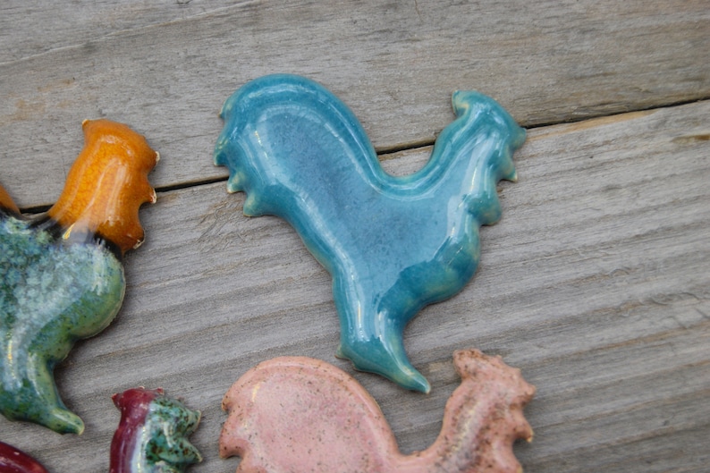 May include: A collection of ceramic rooster figurines in various colors, including teal, orange, pink, and green. The roosters are all in a standing position with their heads tilted to the side.