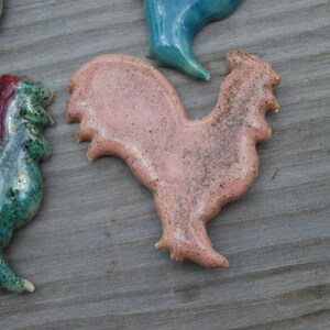 May include: A set of four ceramic rooster figurines. The roosters are in various colors, including pink, green, and blue. The roosters are all in a standing position with their heads tilted to the side.