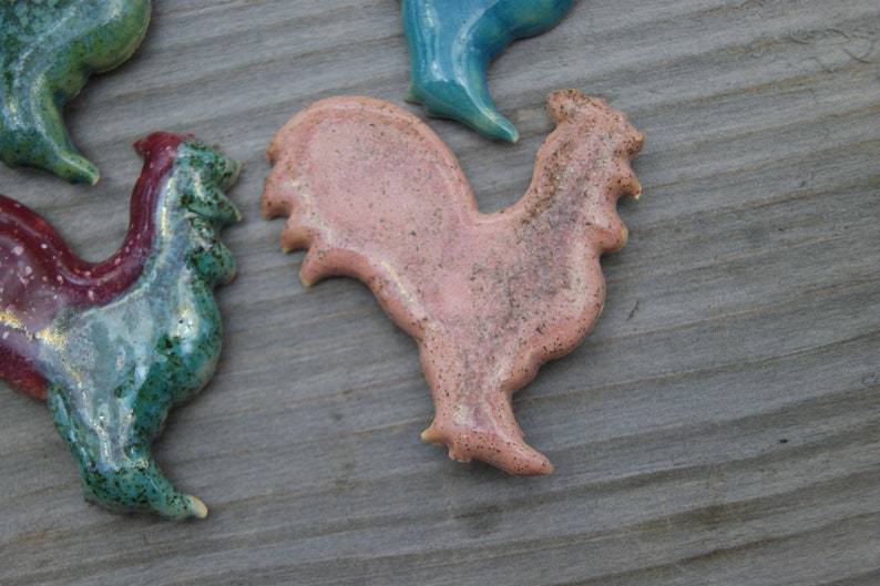 May include: A set of four ceramic rooster figurines. The roosters are in various colors, including pink, green, and blue. The roosters are all in a standing position with their heads tilted to the side.