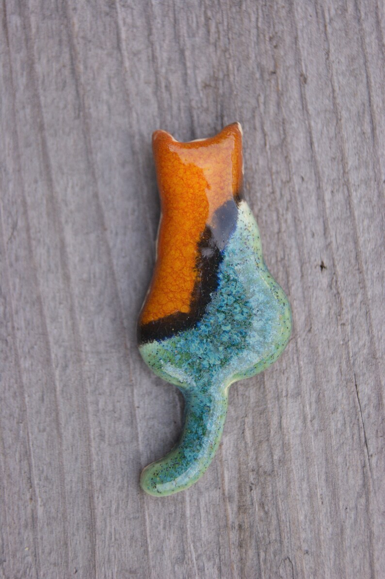 May include: A ceramic cat figurine with a blue, green, and orange glaze. The cat is in a sitting position with its tail curled around its body.