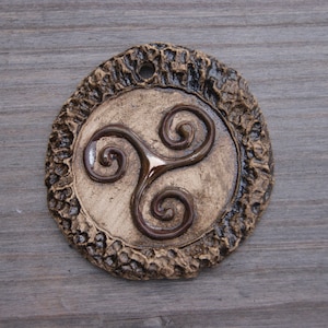 Puede incluir: Un adorno de cerámica marrón con un diseño en espiral de tres partes en relieve. El adorno tiene una superficie rugosa y texturizada.