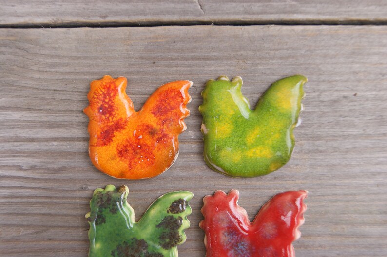 Op de afbeelding: Vier keramische haanfiguren in verschillende kleuren. De hanen zijn oranje met rode vlekken, groen met gele vlekken, rood met zwarte vlekken en groen met bruine vlekken.