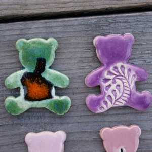 May include: Three ceramic bear figurines. One is green with a black and orange design, one is purple with a white floral design, and one is pink.