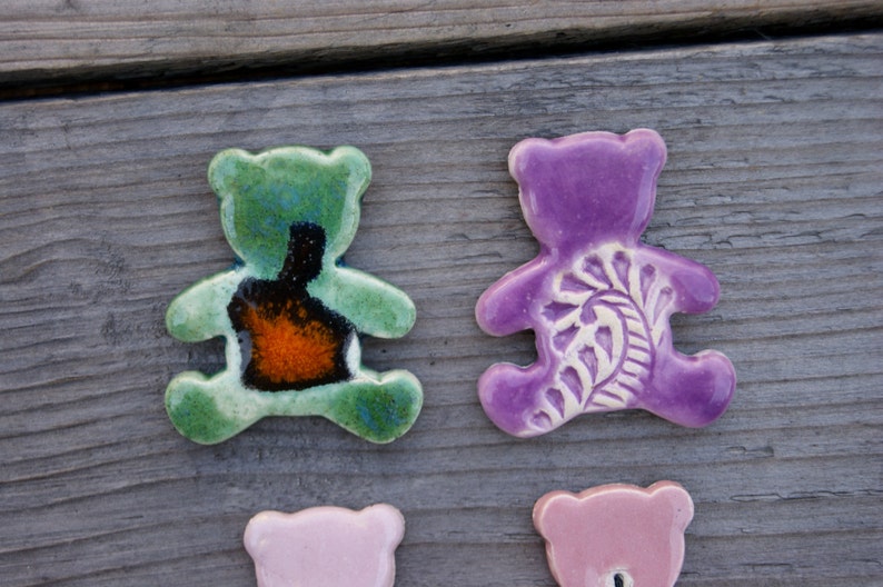 May include: Three ceramic bear figurines. One is green with a black and orange design, one is purple with a white floral design, and one is pink.