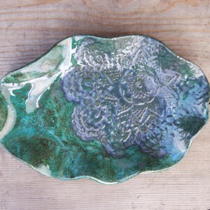 Puede incluir: Un plato de cerámica con un borde ondulado y un esmalte verde y azul. El plato tiene un diseño de flores en relieve.