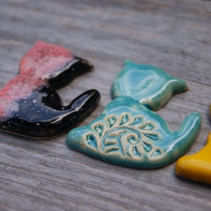 May include: Three ceramic cat figurines in different colors and designs. The first cat is black and pink with a speckled glaze. The second cat is teal blue with a white floral design. The third cat is yellow with green accents.