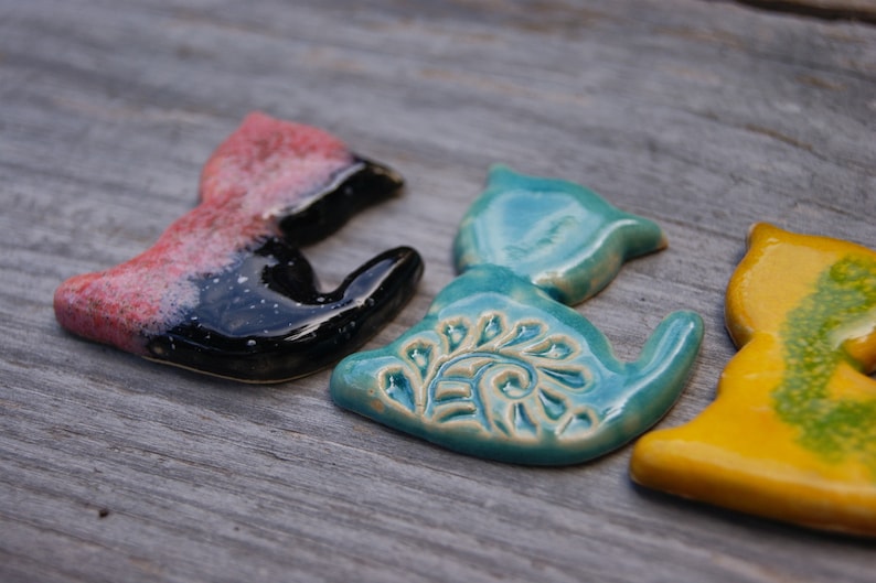 May include: Three ceramic cat figurines in different colors and designs. The first cat is black and pink with a speckled glaze. The second cat is teal blue with a white floral design. The third cat is yellow with green accents.