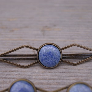 Puede incluir: Una pinza para el pelo azul y bronce con un dise&ntilde;o geom&eacute;trico. La pinza tiene una piedra redonda azul en el centro.