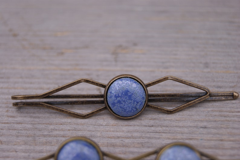 Puede incluir: Una pinza para el pelo azul y bronce con un dise&ntilde;o geom&eacute;trico. La pinza tiene una piedra redonda azul en el centro.