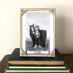 May include: A framed tarot card with a black and white illustration of a person holding a horse's reins. The card is titled "Strength" and the number "VIII" is in the top right corner.