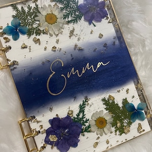 Könnte beinhalten: Ein weißes Notizbuch mit einem blauen und weißen Ombre-Design. Das Notizbuch hat goldene Blatt-Akzente und gepresste Blumen. Der Name "Emma" ist in goldener Schrift auf dem Einband geschrieben.