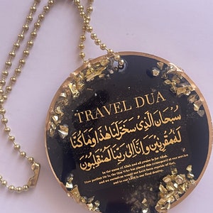 Op de afbeelding: Een zwarte en gouden Travel Dua hanger met gouden tekst en accenten. De hanger heeft een gouden kogelketting en bevat Arabisch schrift en Engelse tekst. De tekst bevat de zin "In the name of Allah and all praise is for Allah."