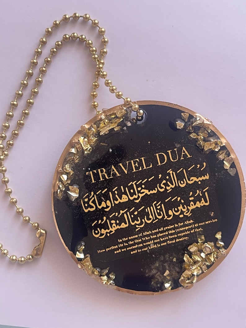 Op de afbeelding: Een zwarte en gouden Travel Dua hanger met gouden tekst en accenten. De hanger heeft een gouden kogelketting en bevat Arabisch schrift en Engelse tekst. De tekst bevat de zin "In the name of Allah and all praise is for Allah."