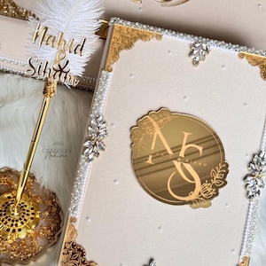 May include: A white guest book with gold accents and a gold mirror with the initials "N & S" surrounded by floral designs. The book is decorated with white pearls and rhinestones. A gold pen with a white feather is in the background.