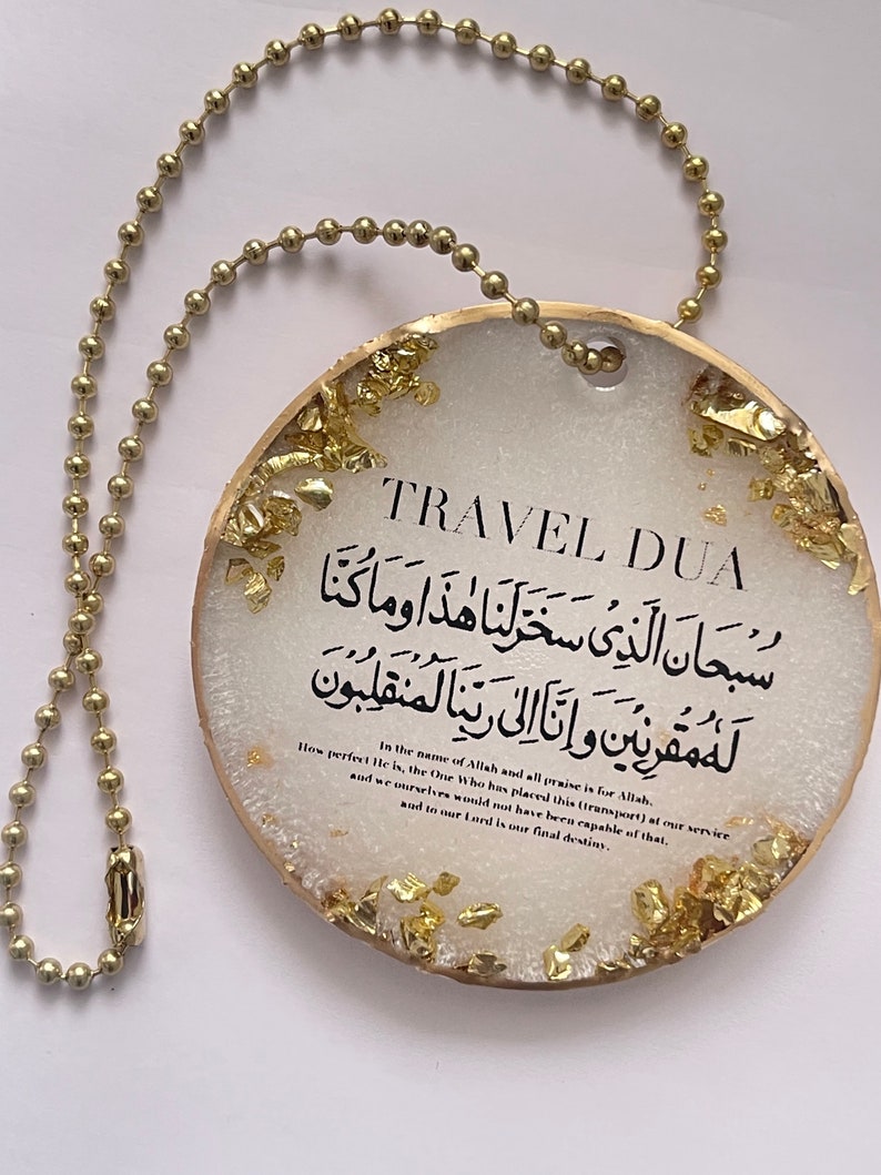 Op de afbeelding: Een rond ornament van wit en goudkleurig hars met de tekst "TRAVEL DUA" en Arabische tekst. Het ornament is afgewerkt met bladgoud en heeft een gouden kogelketting om op te hangen.