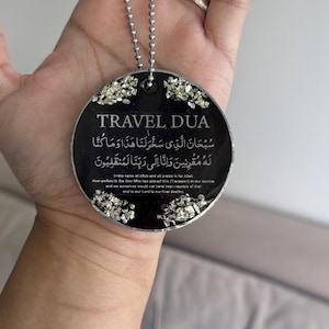 Op de afbeelding: Een zwarte ronde hanger met de tekst "TRAVEL DUA" en Arabisch schrift. De hanger is afgewerkt met zilverkleurige versieringen en hangt aan een zilveren kogelketting.