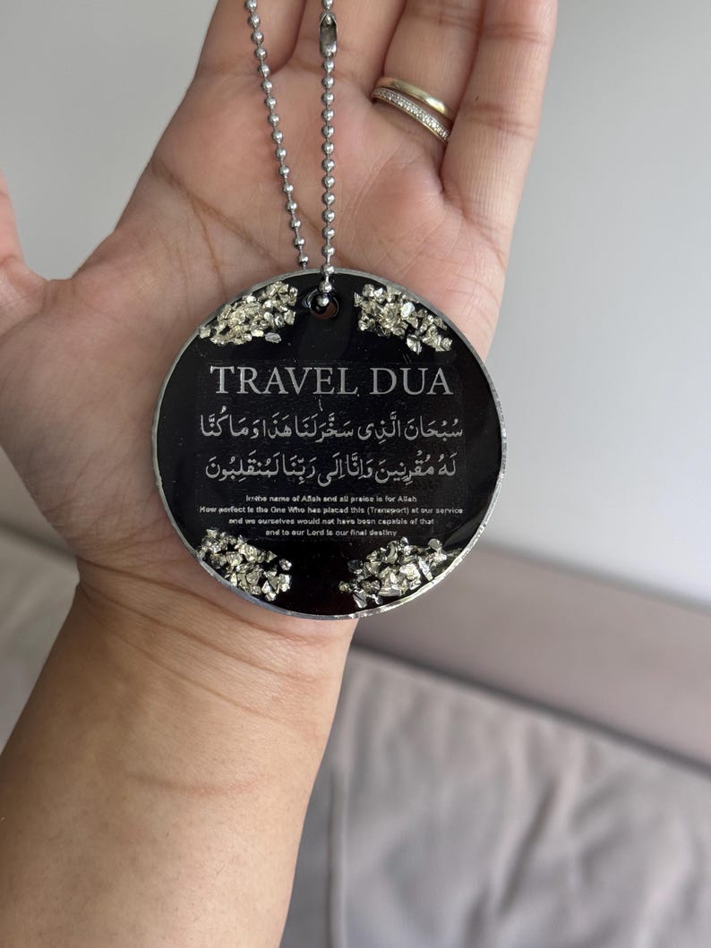 Op de afbeelding: Een zwarte ronde hanger met de tekst "TRAVEL DUA" en Arabisch schrift. De hanger is afgewerkt met zilverkleurige versieringen en hangt aan een zilveren kogelketting.